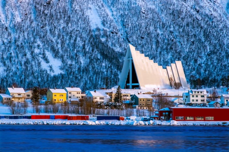 Troms&oslash; city lights glow against the Arctic dusk, framed by snow-covered mountains and the waters of the Troms&oslash;ysundet strait.
